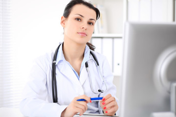 Young brunette female doctor sitting at the table and working by computer  at hospital office.    Doctor is in trouble with diagnosis. Health care, insurance and help concept.