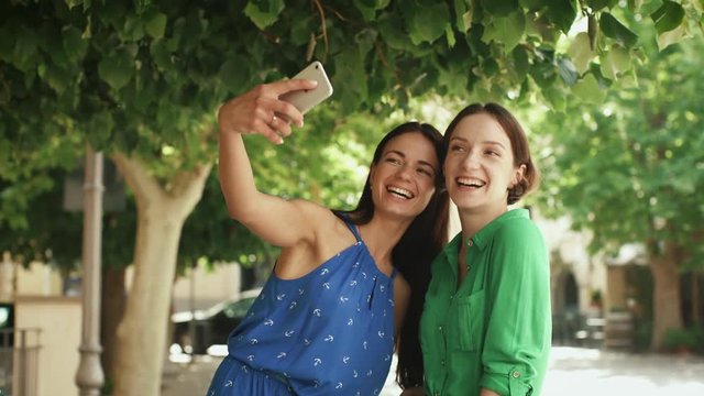 Two Attractive Smiling and Laughing Young Brunette Women in Light Summer Dresses doing Selfies on Streets of European Town.
