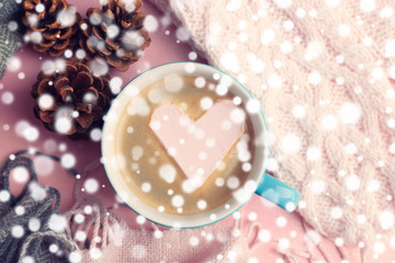 Cup of hot cappuccino with heart marshmallow and warm clothes on pink background, close up. Snow effect