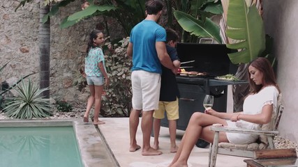 Mother holding wine glass and children enjoying barbecue with father in background - Powered by Adobe