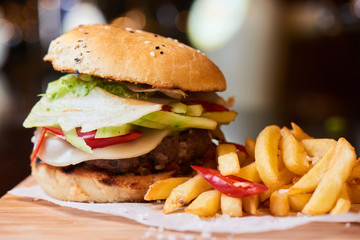 A hamburger consisting of meat patties, vegetables, cheese and French fries served on a wooden plate
