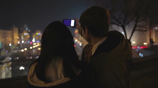 Young couple in love taking selfie and kissing at romantic date, night cityscape