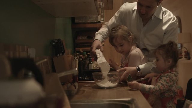 Father and daughters making pancake in kitchen, father pouring flour into mixing bowl