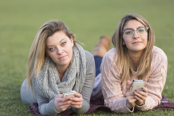 Portrait of two friends lying down on their stomach on grass holding smartphone