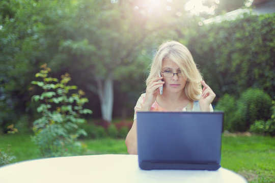 Красивая женщина сидит за столом в саду с ноутбуком и мобильным телефоном