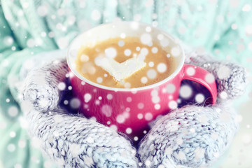 Female hands in warm mittens holding cup of hot cappuccino with heart marshmallow, close up. Snow effect