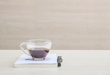 Closeup black coffee in transparent cup of coffee on white book with black pen on blurred wooden desk and wall textured background in the meeting room under window , relaxation in coffee break concept