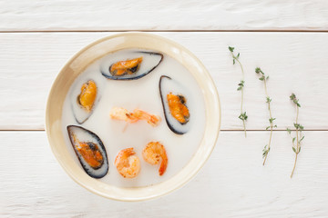 Bowl of seafood cream-soup with rosemary flat lay. Top view on bowl with white cream-soup with mussels and shrimps on white wooden background, decorated with rosemary, void. Creative food decoration