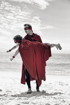 Father in superhero costume lifting son at sea shore