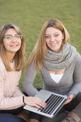 Smiling young women sitting on the grass with legs crossed, holding a portable computer and looking at the camera