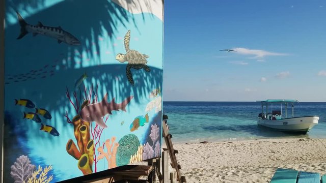 Silk Caye, Belize - March, 2016 - Static shot of a mural shaded by palm trees in the foreground and a bird and a boat in the background.
