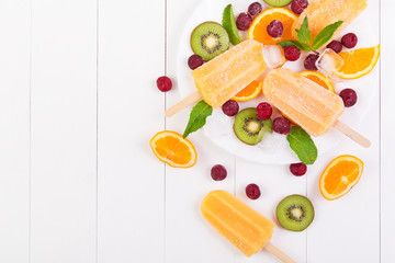 Homemade orange popsicles on a plate with mint, kiwi, frozen cherry and orange slices on a white wooden table. Top view with copyspace