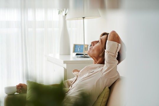 Mature woman sitting on sofa, contemplating and drinking tea