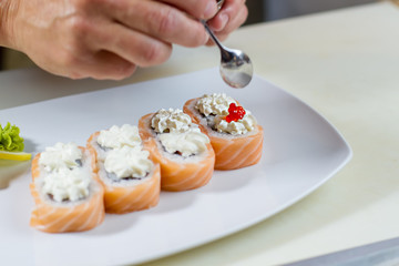 Hand holds spoon over sushi. Red caviar on sushi roll. Salmon caviar for uramaki rolls. Delicacy from japanese cuisine.