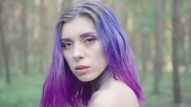 Close up of pretty woman face shaking her purple hair and looking into the camera. Isolated in forest, in slow motion.
