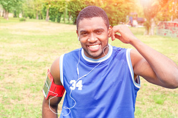 Naklejka premium African american man jogging at city park - Black runner gay listening music with earphones while training outdoor - Concept of healthy lifestyle and sport activity with toothy smile positive person