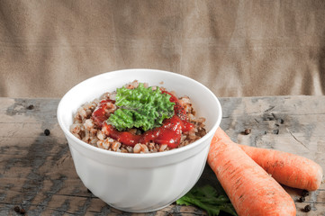 Buckwheat porridge with tomato sauce and leaf salad in a white bowl on a wooden table next to a ceramic bowl with dish is carrots or sliced carrot. Food background. Nice and tasty atmosphere.
