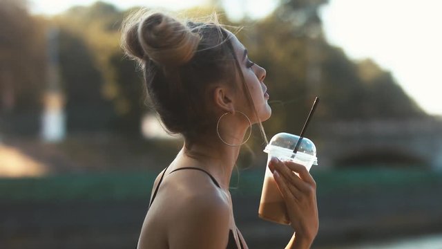 Urban portrait of young attractive blonde girl in jeans drinking milkshake in the park. 60 FPS slow motion, 4K UHD RAW color graded footage