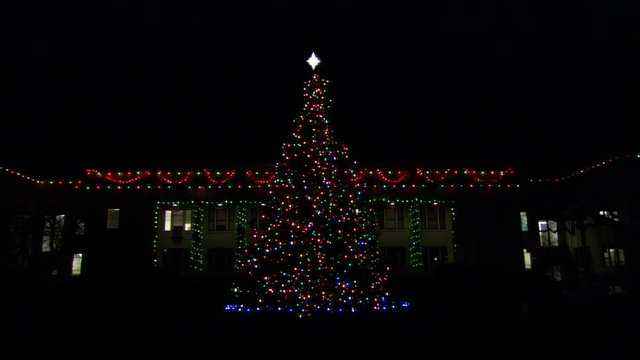 An outdoor Christmas tree in front of a large house trimmed with lights at night