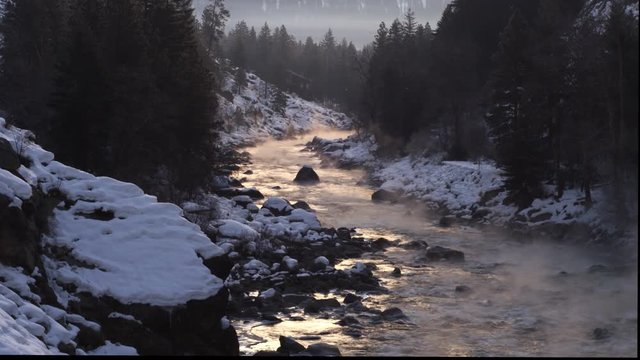 Evening light on a misty river flowing through a snowy forest canyon, secluded lodge in background