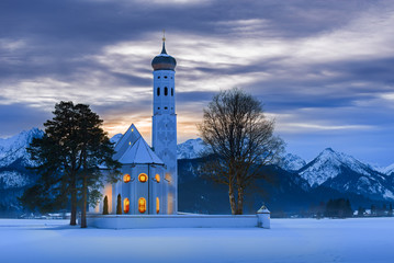 St. Coloman church at dusk on a winter Christmas night with snow and fog and warm candlelight inside