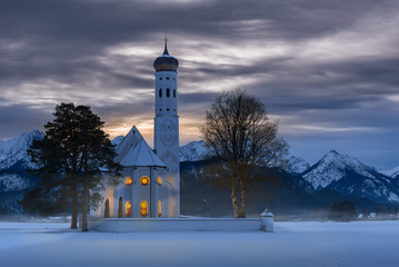 The exterior of the St. Coloman church at dusk on a wintry Christmas evening with warm light coming...