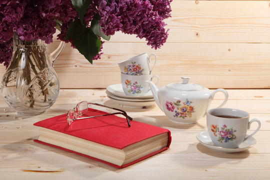 Bouquet of lilacs, book, spectacles, teapot and cup of tea on wo