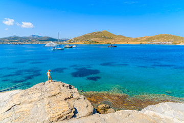 Fototapeta na wymiar Young woman tourist standing on a rock and looking at beautiful Monastiri bay with turquoise sea water, Paros island, Greece