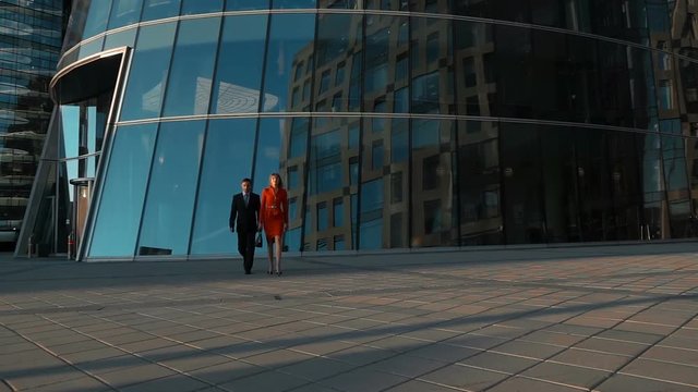 Slow motion. Two successful young businesspeople walk with glass modern building at bg. Businessman and businesswoman seriously winner teamwork concept. Wide shot. Teal and orange.
