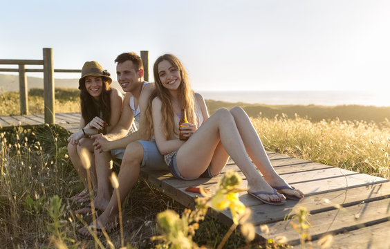Friends sitting on boardwalk at the beach, drinking beer at sunset