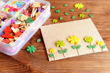 Fototapeta na wymiar Summer card with yellow and green flowers. Postcard made of cardboard, buttons and cord. A box of colorful buttons on a brown wooden background. Simple kids crafts