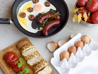 Fried sausages and eggs in a pan served with bread, tomatoes and egg shell on a grey stone board