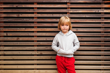 Fashion portrait of adorable toddler boy wearing grey sweatshirt and red trainings, standing against wooden background