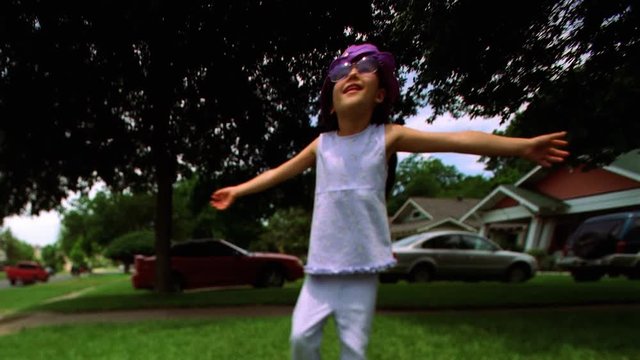 Little girl in fake sunglasses and a purple hat spinning around on a green lawn