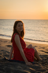 Fototapeta na wymiar Girl in a red dress at sunrise on a golden beach watching the sea silhouette