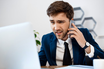 Annoyed young businessman speaking on phone, office background.