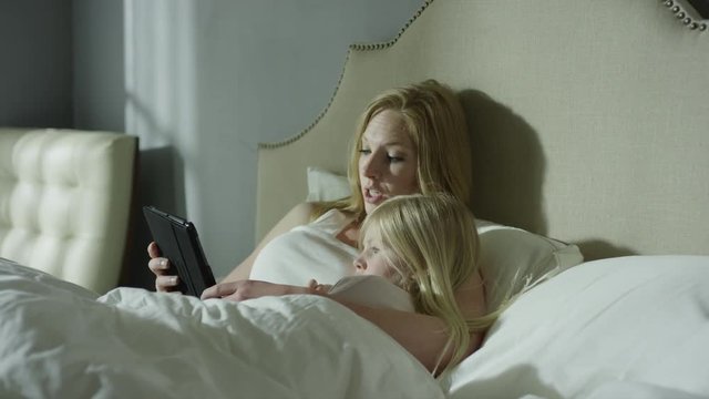 Medium panning shot of mother reading e-book to daughter in bed / Cedar Hills, Utah, United States