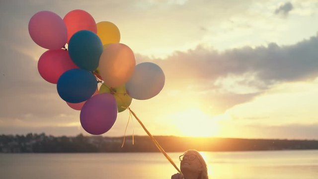 Young woman with helium balloons in hand walking on the beach at sunset, concept about birthday and happy events. RAW video record.