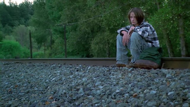 Weary teenage boy sitting on railroad tracks with backpack beside him