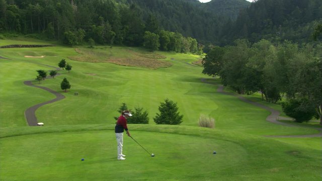 Woman golfer teeing off from hilltop tee block, seen from above at a slight distance