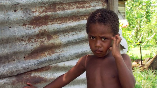 Little Fijian boy standing by corrugated metal wall and scowling at camera