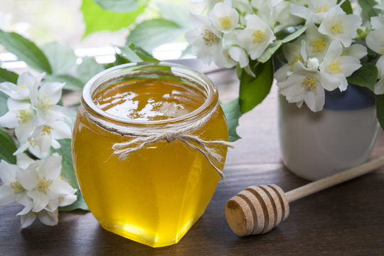 Delicious fresh spring honey in glass jar, wooden spoon and blooming branch and flowers of jasmine in white-blue ceramic cap on black stone board against  background of window