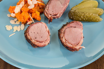 Drei Scheiben gegrilltes Schweinefilet im Speckmantel, angerichtet auf blauem Teller mit Karottengemüse, garniert mit Gewürzgurke und Mandelblättern