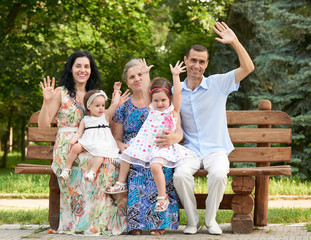 Fototapeta premium big family sit on wooden bench in city park and waving, summer season, child, parent and grandmother, group of five people
