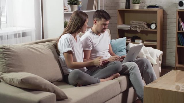 Young boyfriend and girlfriend sitting on the couch and using laptop to make online order with credit card 