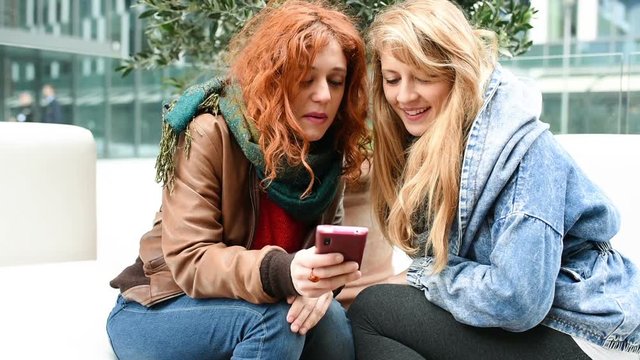 Two young beautiful caucasian blonde and redhead woman sitting outdoor chatting and laughing, looking the screen of a smart phone hand hold -  communication, friendship, technology concept