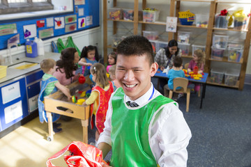 Teacher in a classroom. He is holding some aprons and smiling at the camera. The children are playing in the background with other teachers.
