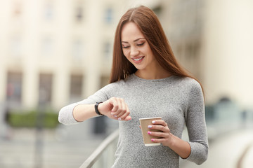 Time to work! Coffee break. Beautiful young woman holding paper cup and looking for wrist watch. Business concept.