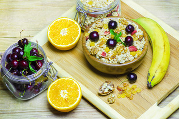 Healthy breakfast ingredients.Bowl of oat granola with cherries and nuts,fresh cherries in open glass jar,dry fruits,banana and oranges on wooden board