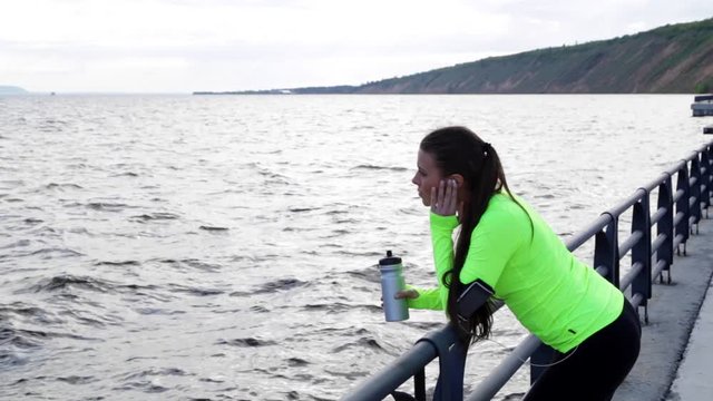 Sport woman runner jogger look by sea. She is exercising and take a break and holding bottle of water on city embankment. View of mountain range is in background.